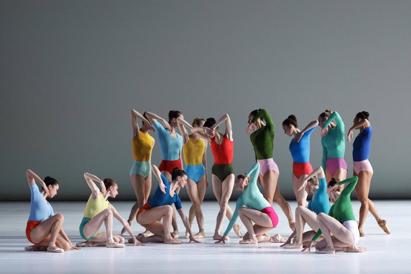 a corps of female dancers pose in profile, all with one elbow sharply pointing up towards the sky, some stand others are seated,  they wear tight color-ful costumes in two bright tones, one bright tone on the top and another on the bottom, a feast of color
